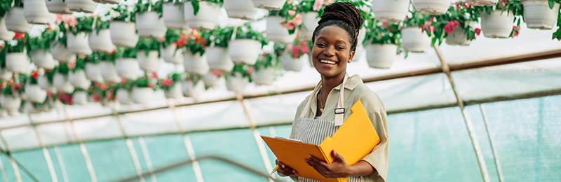 Female entrepreneur standing in a greenhouse with hanging flower pots holding a folder that includes additional data and charts about her business.